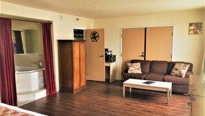 Een kamer met een bed, een houten garderobe, een bruine bank met gestreepte kussens, een witte salontafel en een klein keukenhoekje met een magnetron.