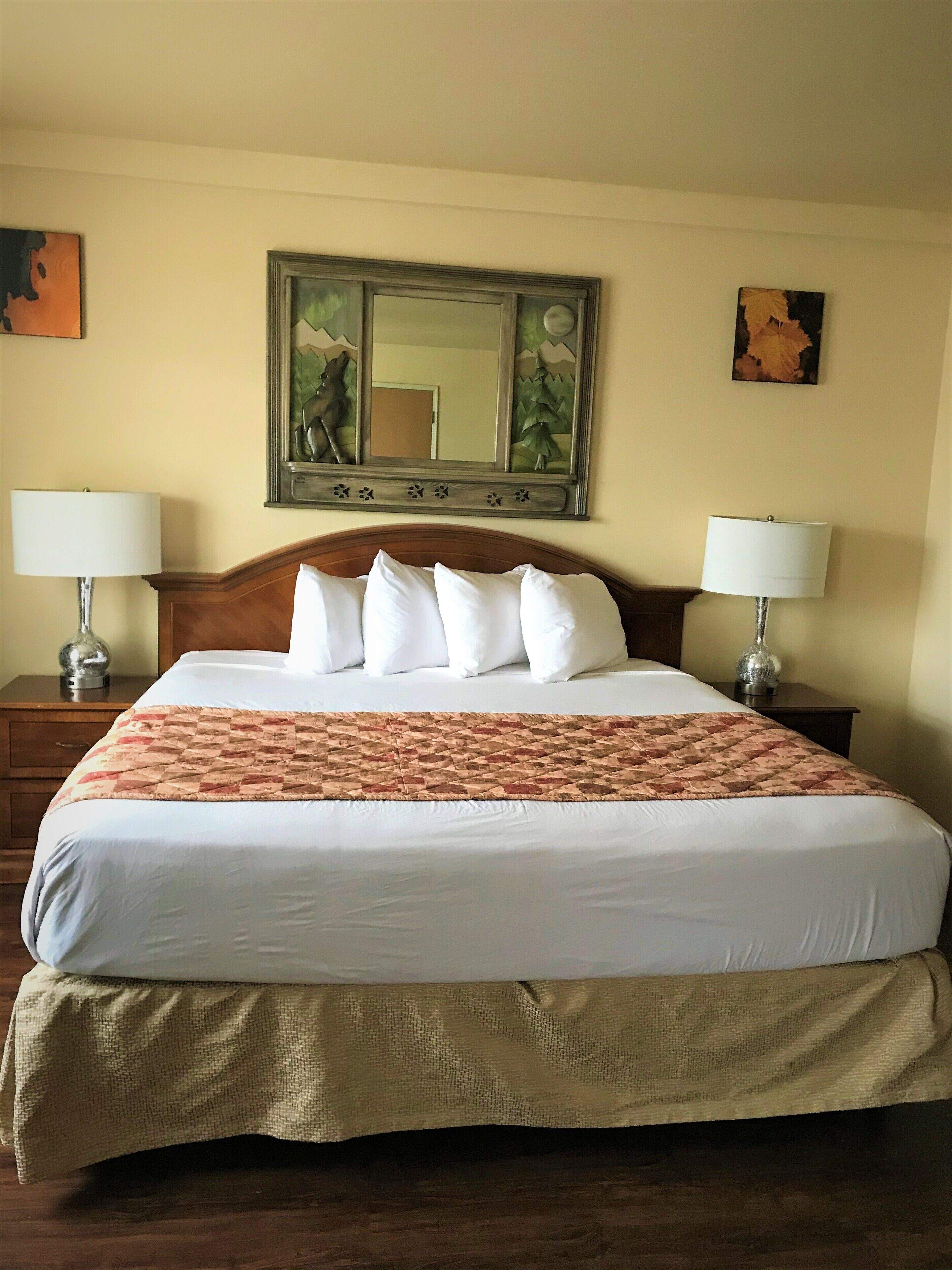 Una cama bien hecha con sábanas blancas y una manta estampada, flanqueada por dos mesitas de noche con lámparas, en una habitación con un espejo grande sobre la cama y obras de arte en las paredes.
