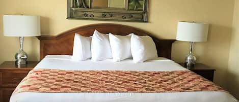 Una cama bien hecha con sábanas blancas y una manta estampada, flanqueada por dos mesitas de noche con lámparas, en una habitación con un espejo grande sobre la cama y obras de arte en las paredes.