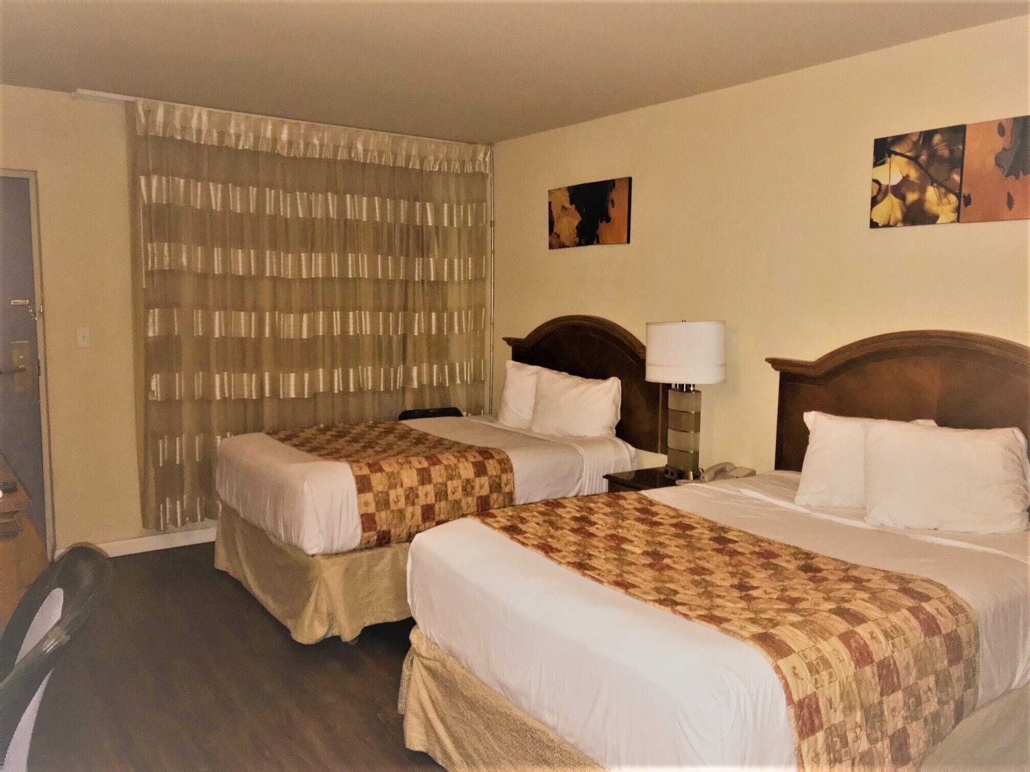 A hotel room with two beds, a wooden headboard, a nightstand, a lamp, and a curtain.
