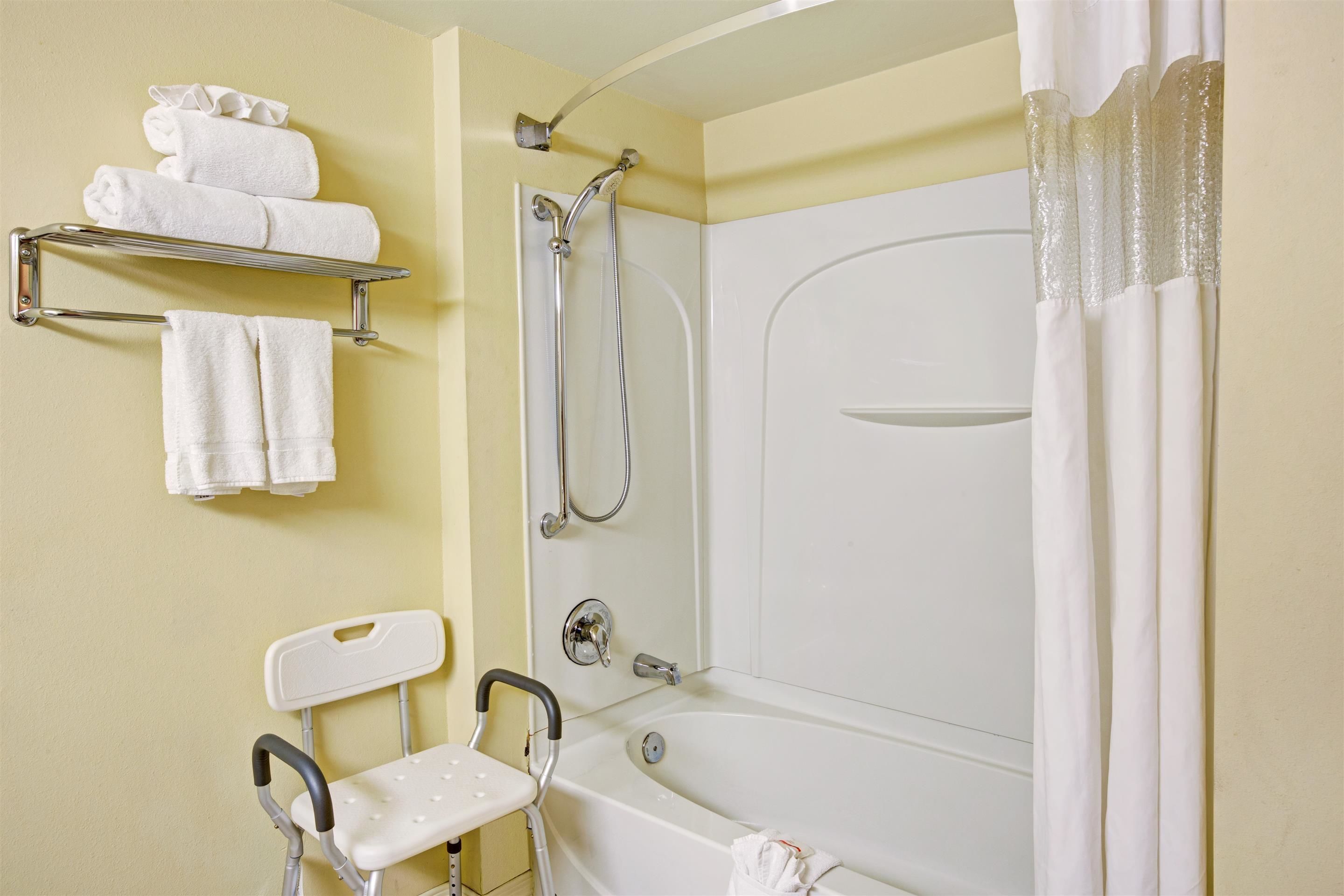 A bathroom with a shower, a white bathtub, a white chair, and a towel rack with folded towels.