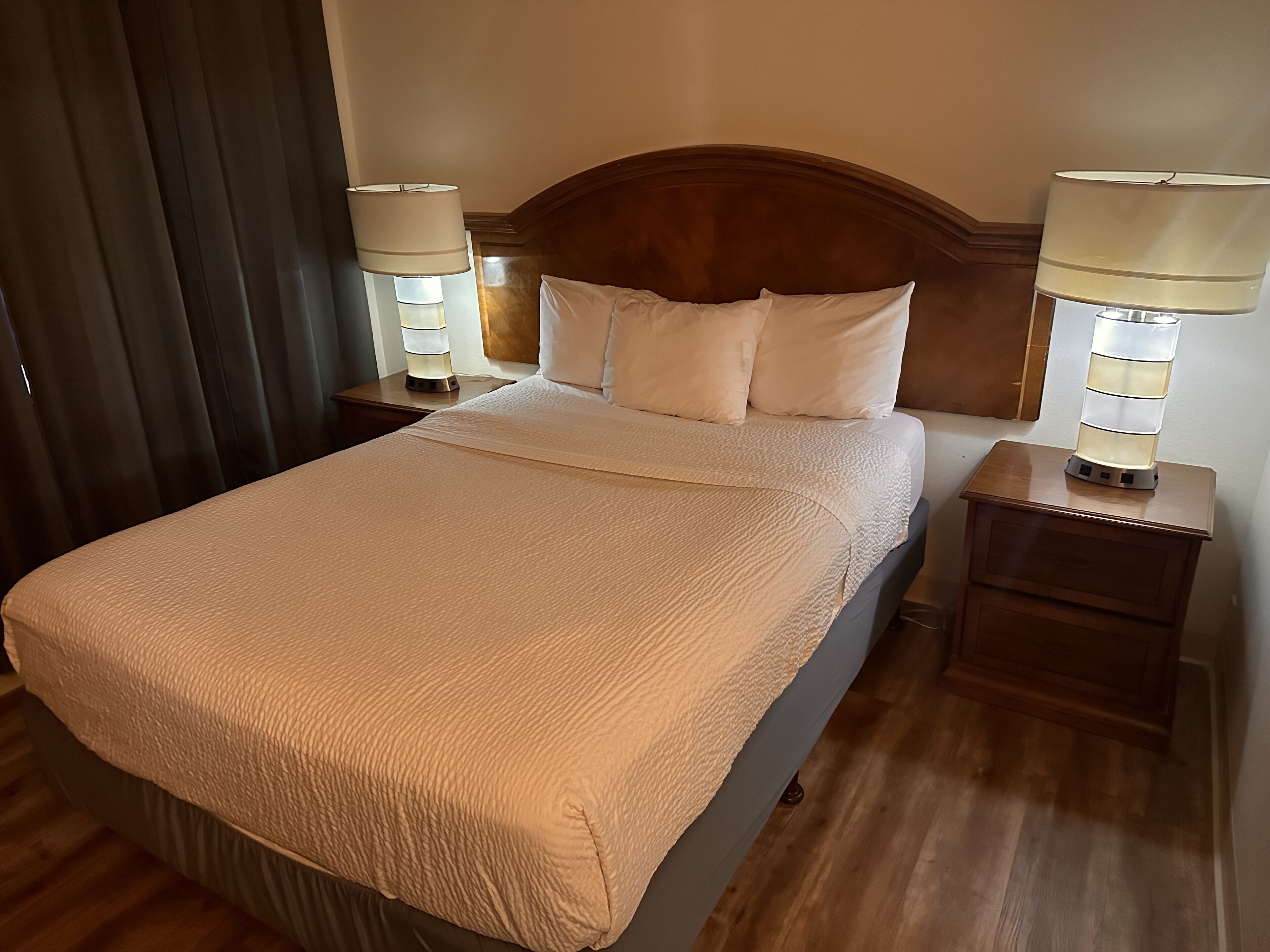 Uma cama bem arrumada com cobertura bege texturizada, ladeada por duas mesas de cabeceira com abajures.
