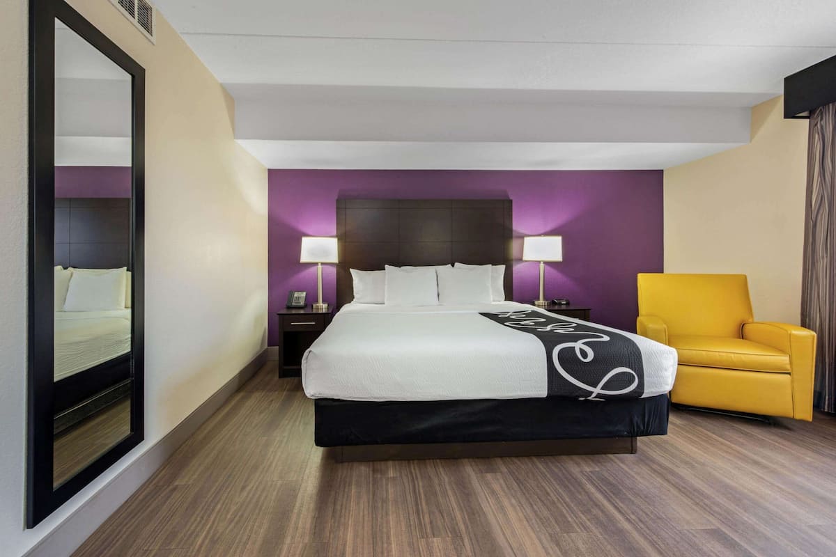 A hotel room with a large bed, a nightstand with a lamp, a mirror, and a yellow armchair.