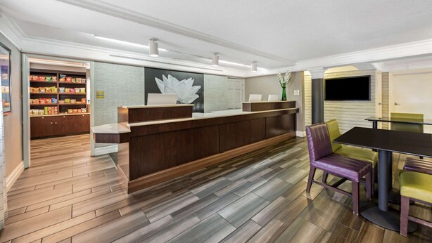 Ein moderner Empfangsbereich mit Holztresen, einer Sitzecke mit violetten und grünen Stühlen und einer im Hintergrund sichtbaren Küche.
