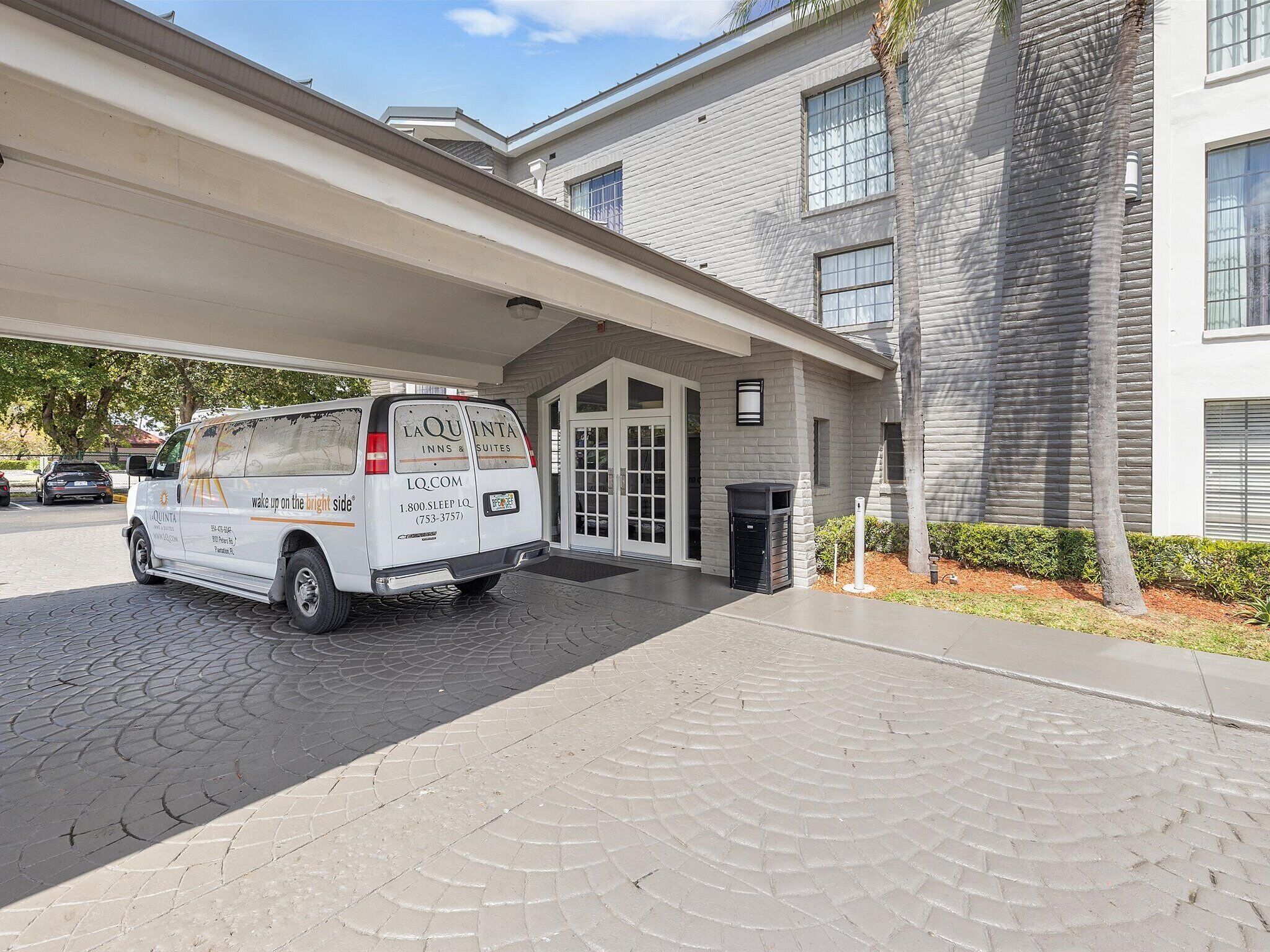 A white van parked in front of a building with a covered entrance and a palm tree nearby.