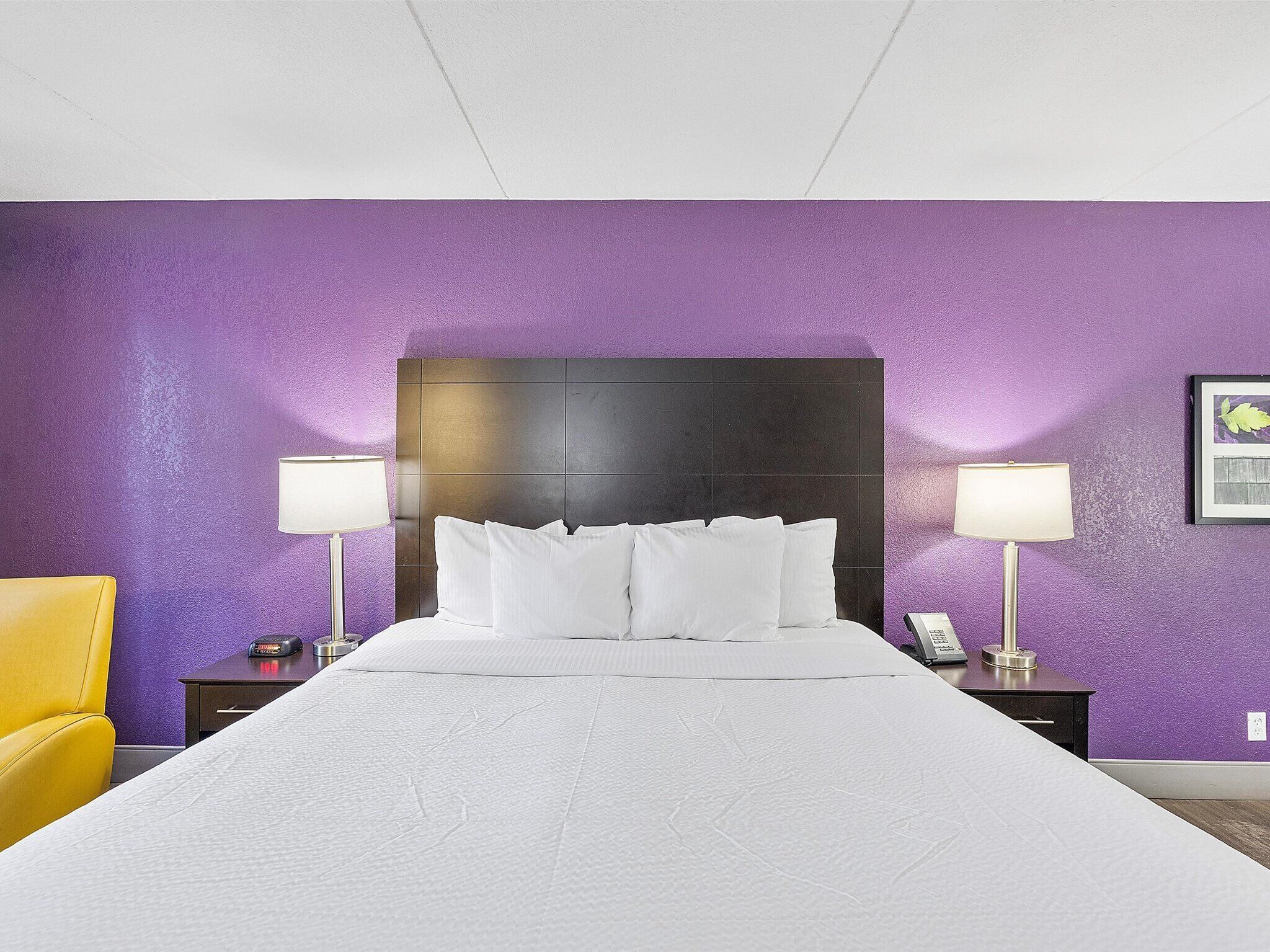 A hotel room with a large bed, two bedside tables with lamps, a chair, and a purple wall.