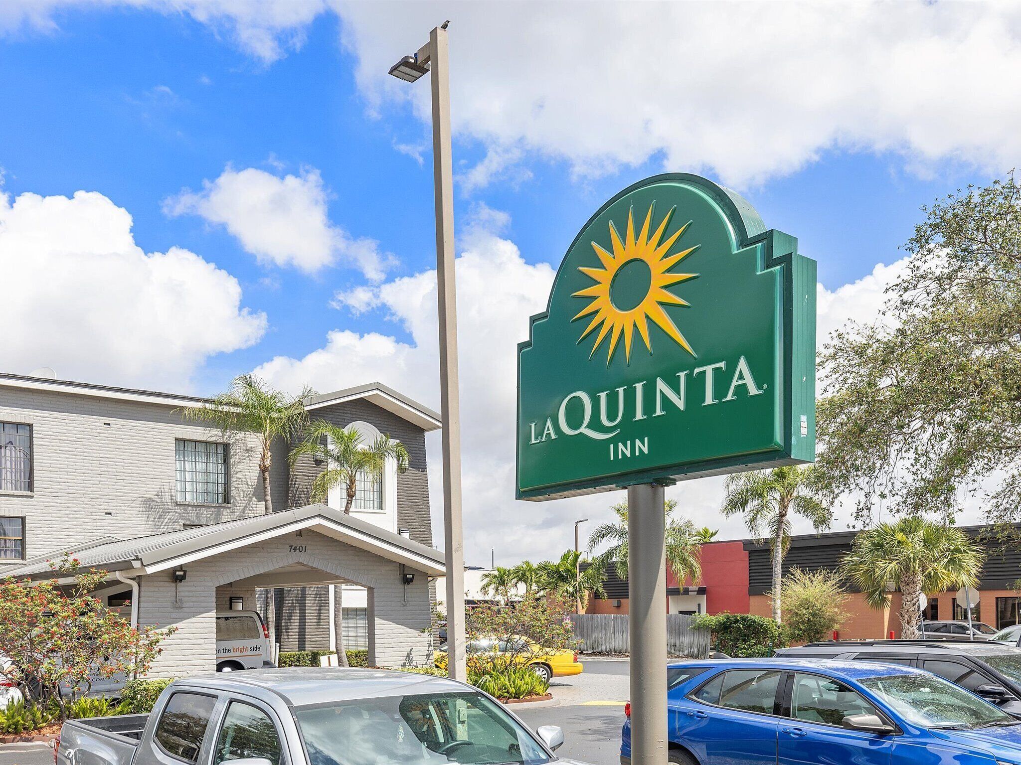 A La Quinta Inn sign with a sun logo, a building with a flat roof and a covered entrance, and several parked cars.