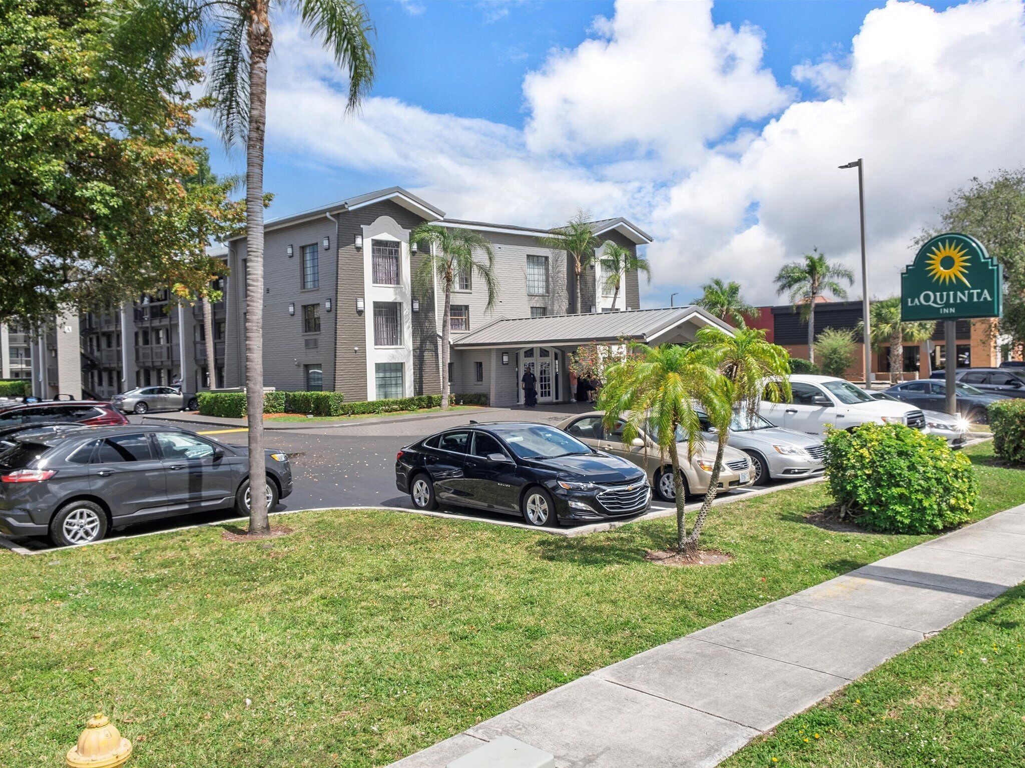 A hotel with a modern design, featuring a parking lot with several cars, a grassy area with palm trees, and a clear sky.