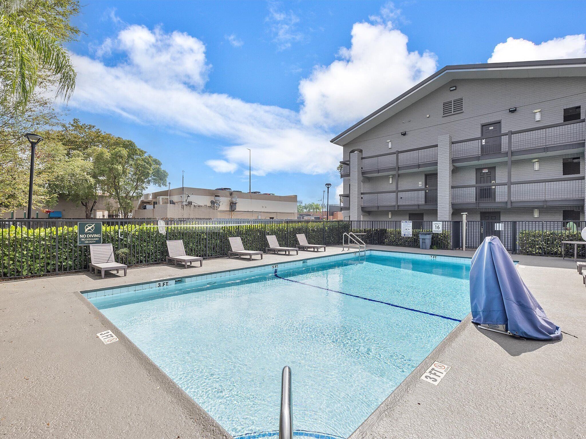 A rectangular swimming pool with a 5 ft depth marker, surrounded by lounge chairs and a covered umbrella.