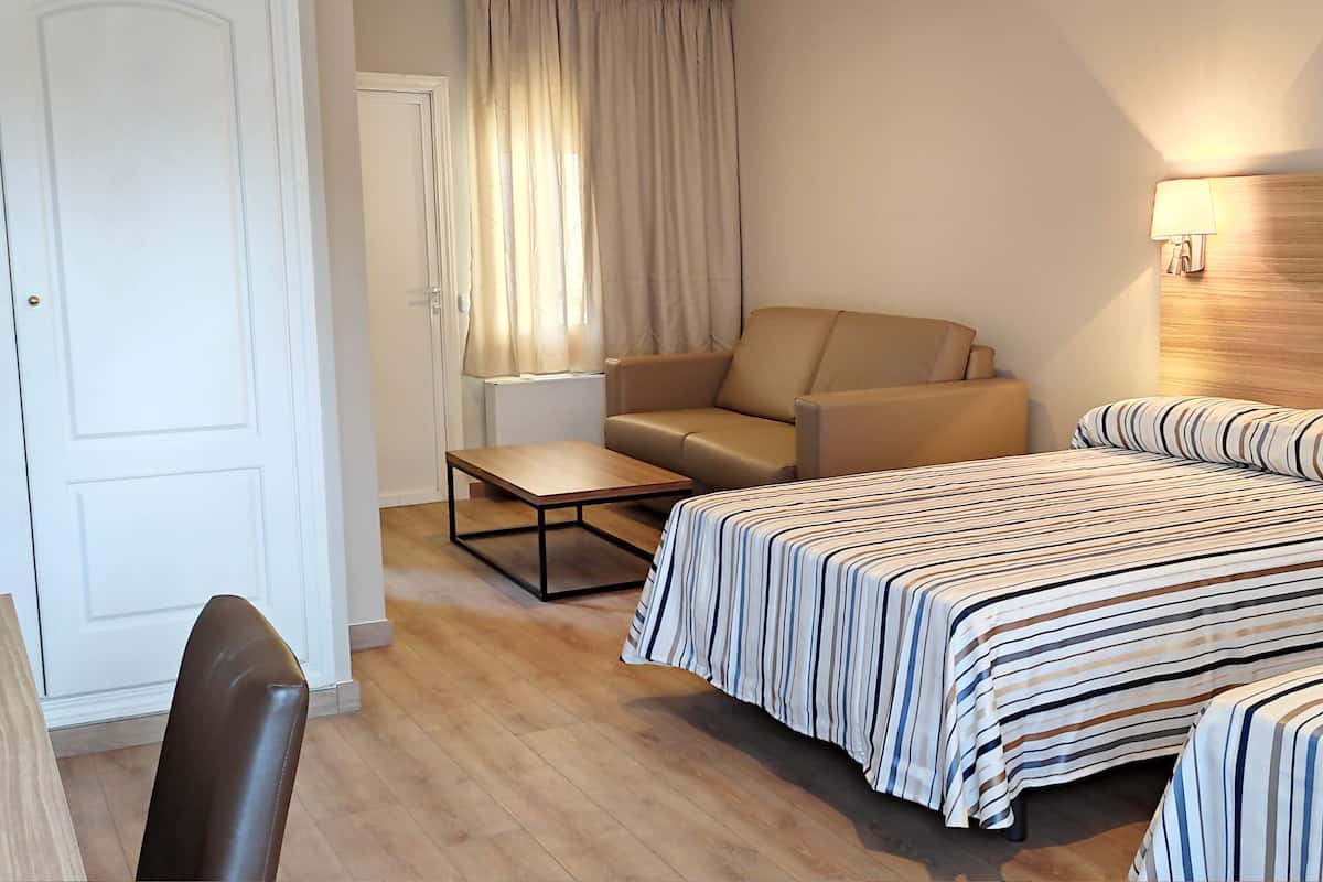 Quarto de hotel com cama, uma cadeira, uma mesinha e janela com cortinas.