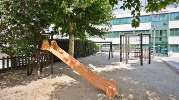 Een speeltuin met een glijbaan en klimconstructies, omringd door bomen en een gebouw op de achtergrond.