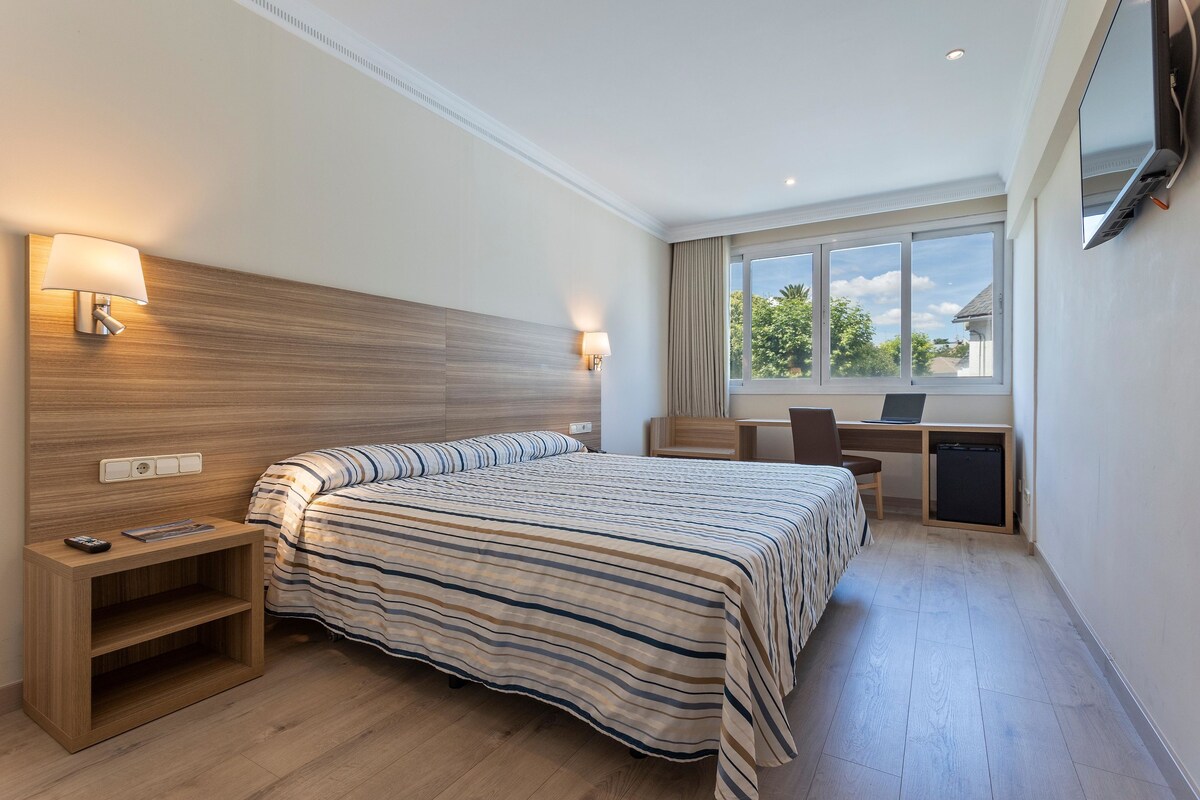 A hotel room with a bed, bedside tables, a desk with a chair, a TV mounted on the wall, and a window with a view of greenery.