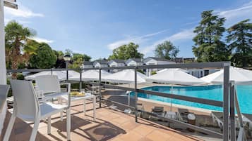 Zona junto a la piscina con sillas y sombrillas blancas, una piscina y vistas a árboles y edificios.