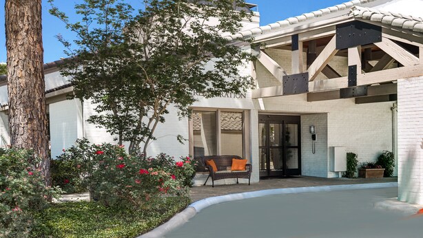 A modern building with a white exterior, a covered patio area with a wicker sofa, and a tree in the foreground. - Value Suites Extended Stay - Arlington (Arlington)
