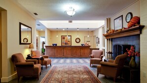 A spacious living room with a large rug, two armchairs, a sofa, a fireplace, and a wooden reception desk in the background. - Value Suites Extended Stay - Arlington (Arlington)