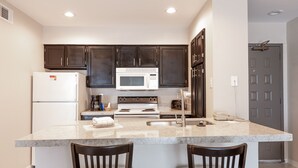 A compact kitchen with dark cabinets, a white refrigerator, a microwave, and a stove. There are two bar stools at the island. - Value Suites Extended Stay - Arlington (Arlington)