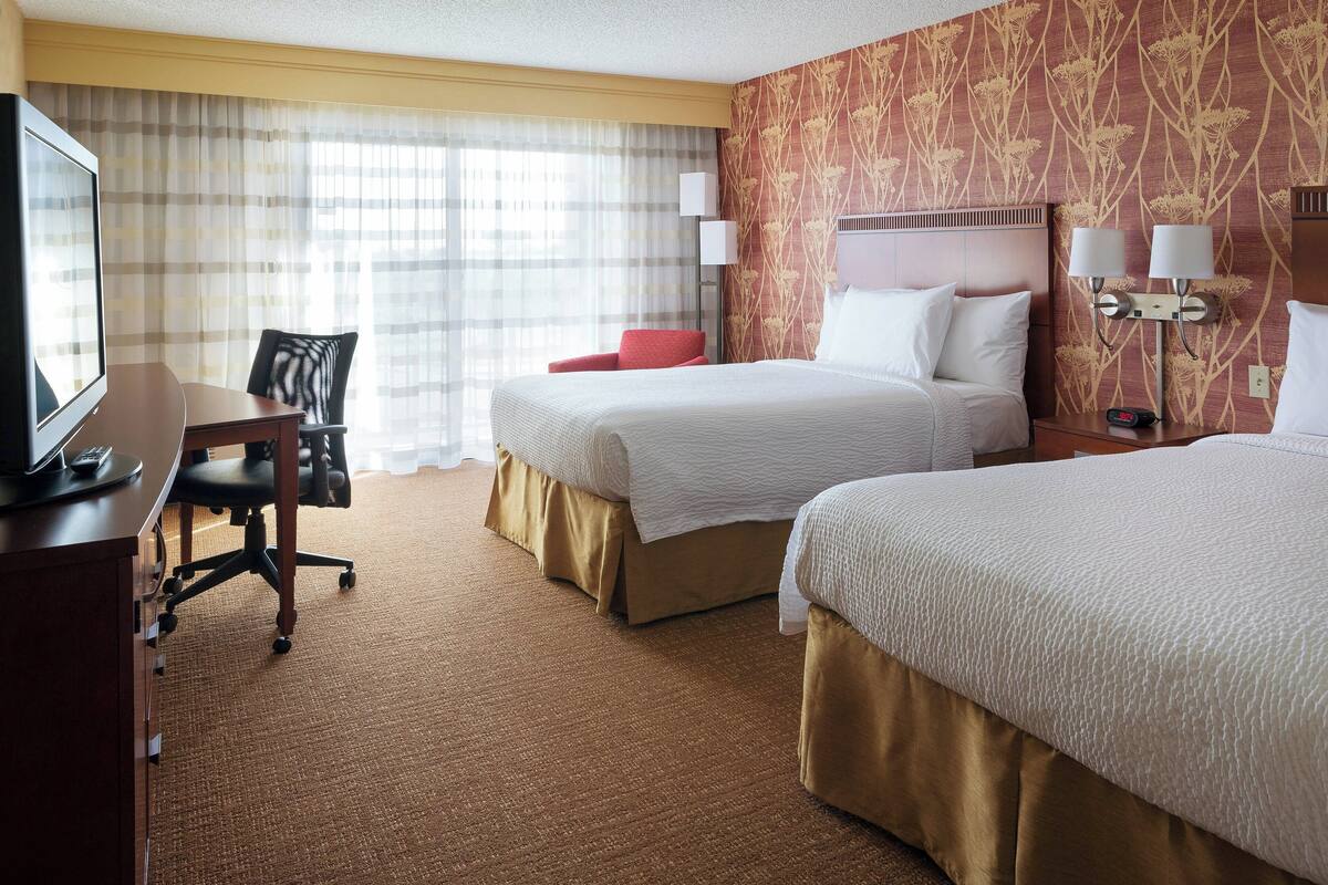 A hotel room with two beds, a desk with a chair, a TV, and a large window with curtains.