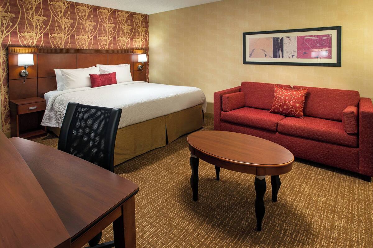 A hotel room with a bed, a red sofa, a wooden table, and a carpeted floor.