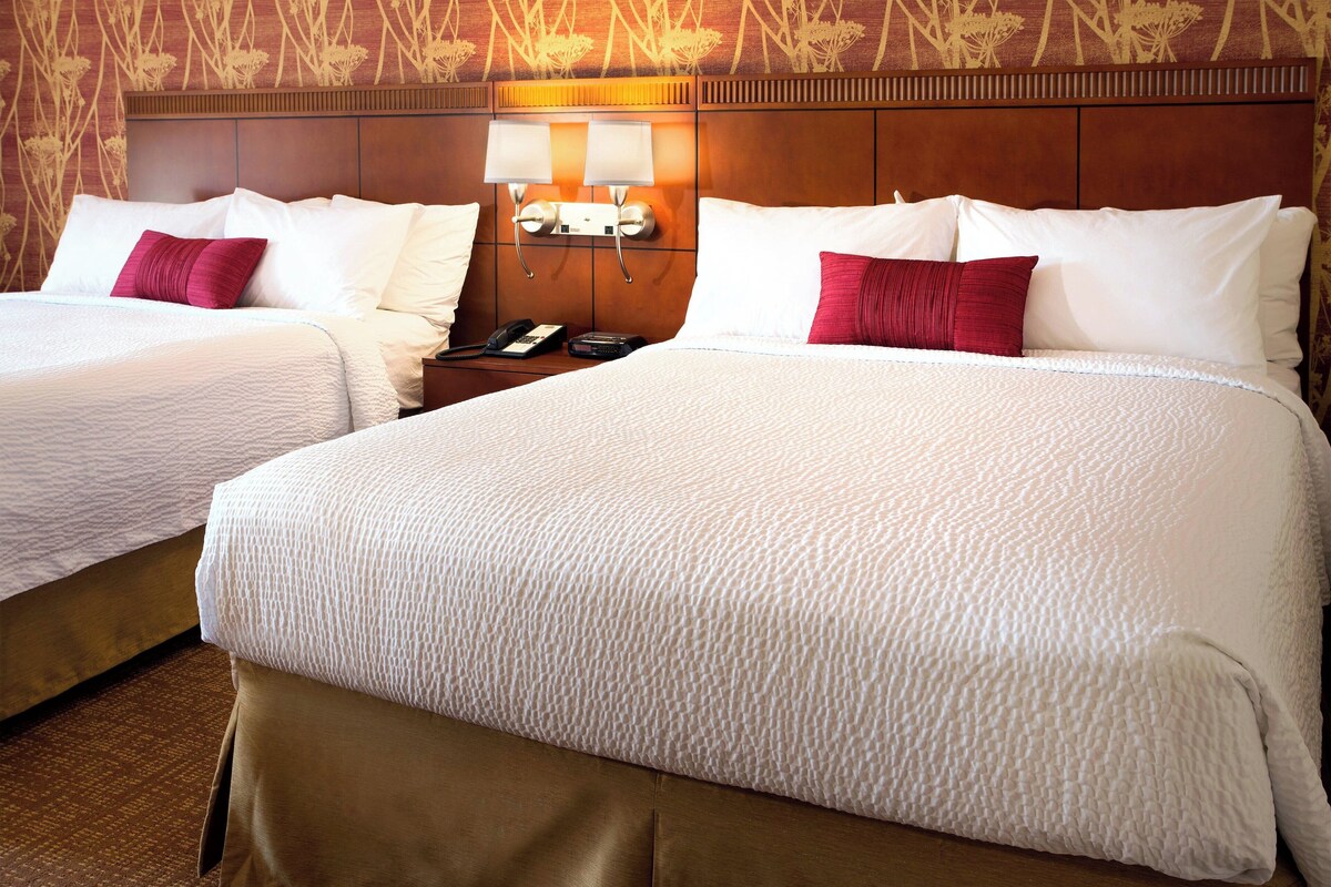 A hotel room with two beds, each with white bedding and red pillows, a wooden headboard, and a nightstand with a phone and remote control.