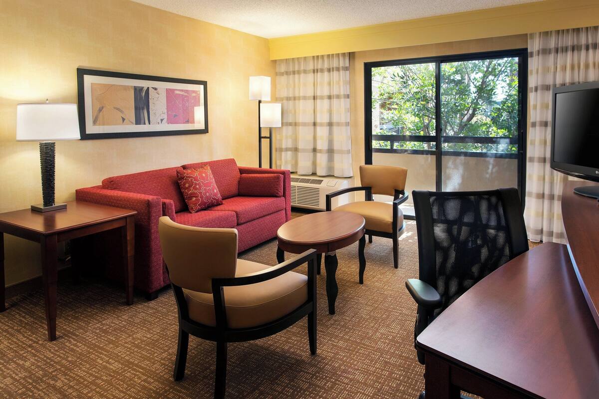 A hotel room with a red sofa, beige armchairs, a small round table, a TV, and a large window with a view of trees.