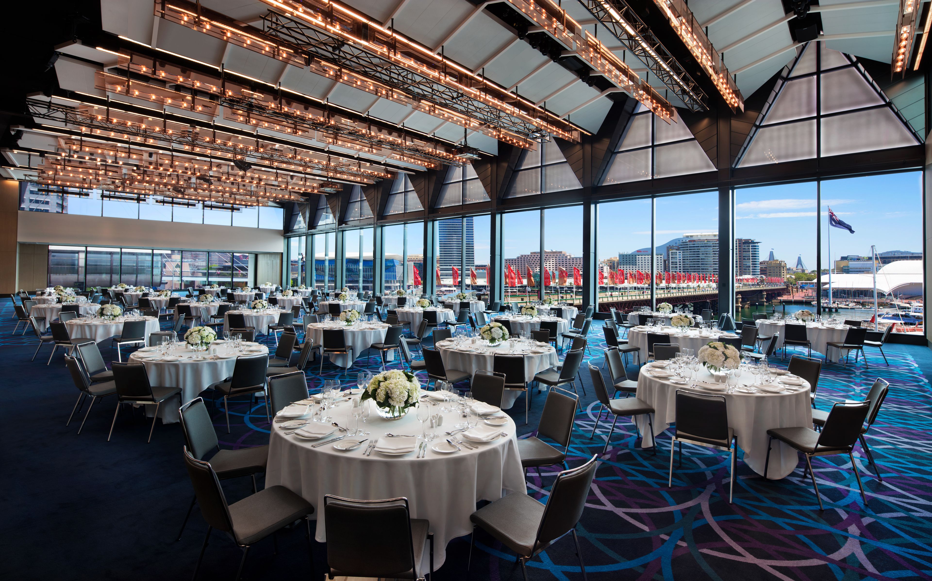 A large, well-lit banquet hall with round tables set for dining, featuring a panoramic city view through large windows.