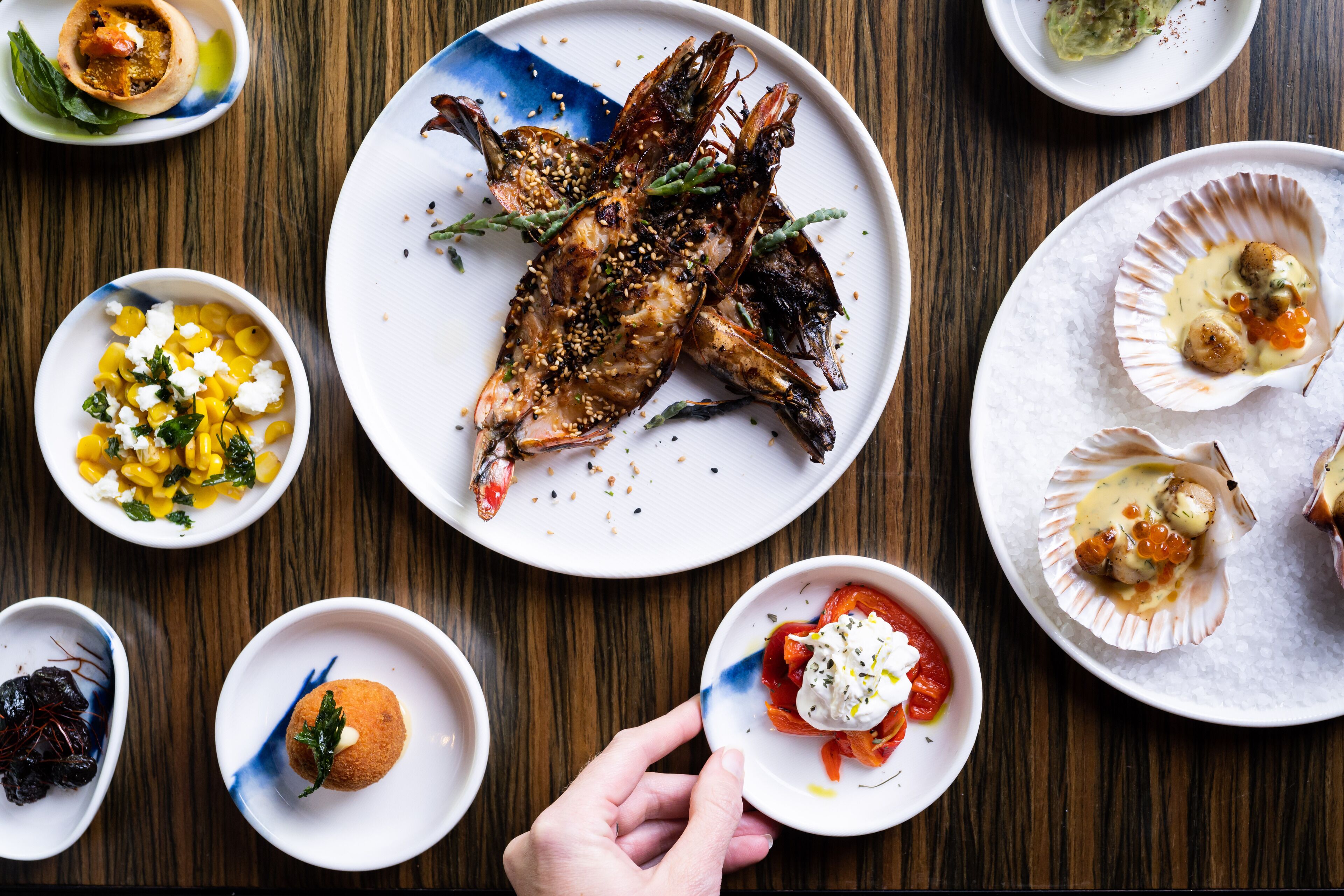A variety of dishes on a wooden table, including grilled seafood, corn salad, and a dessert.