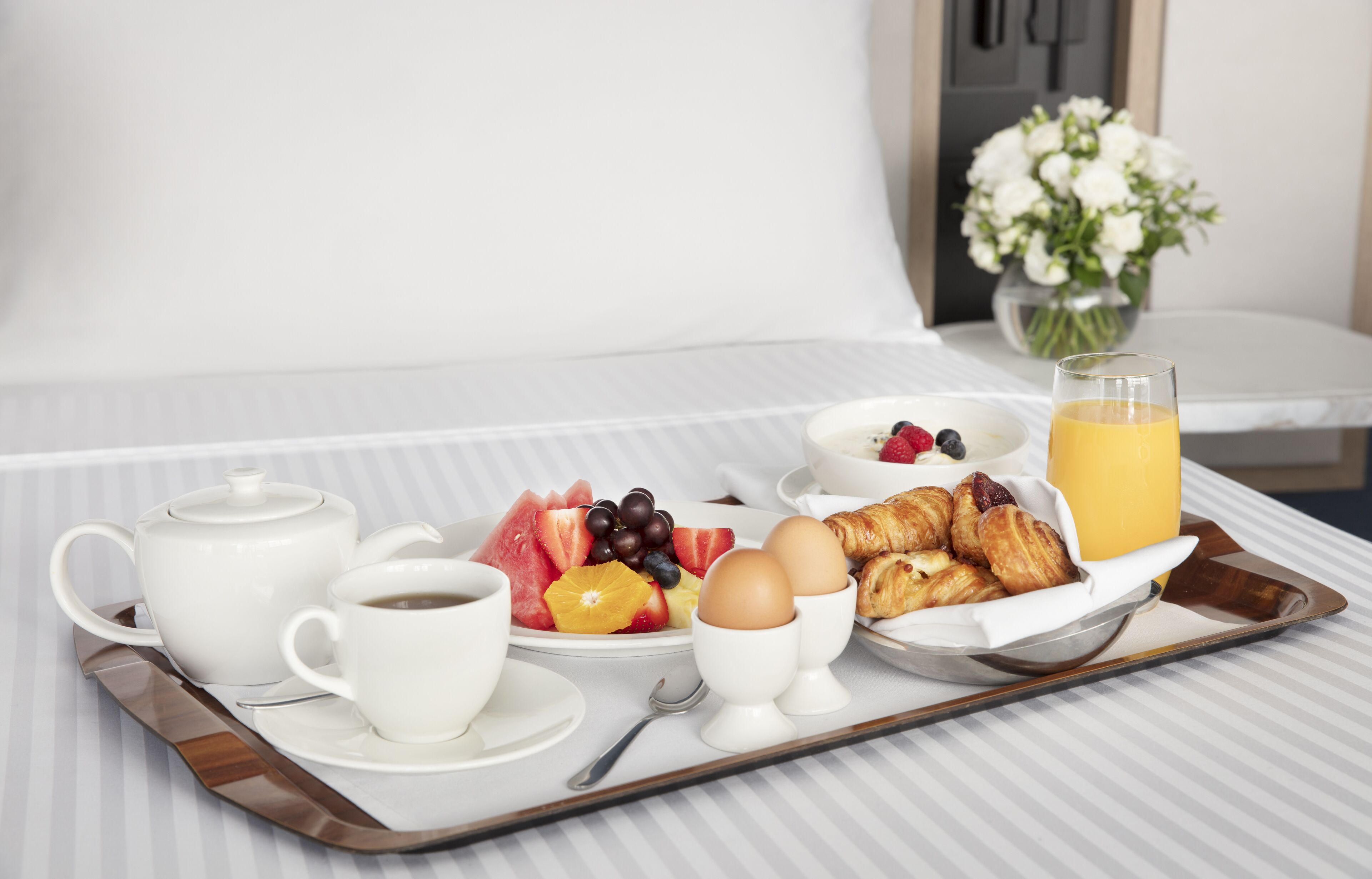 A breakfast tray with croissants, eggs, fruit, and juice on a hotel bed.