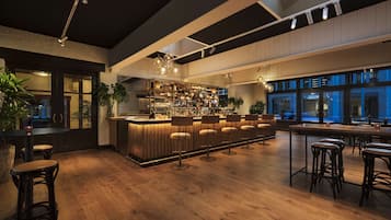 Una zona de bar con suelo de madera, taburetes y una barra bien iluminada con estantes llenos de botellas.
