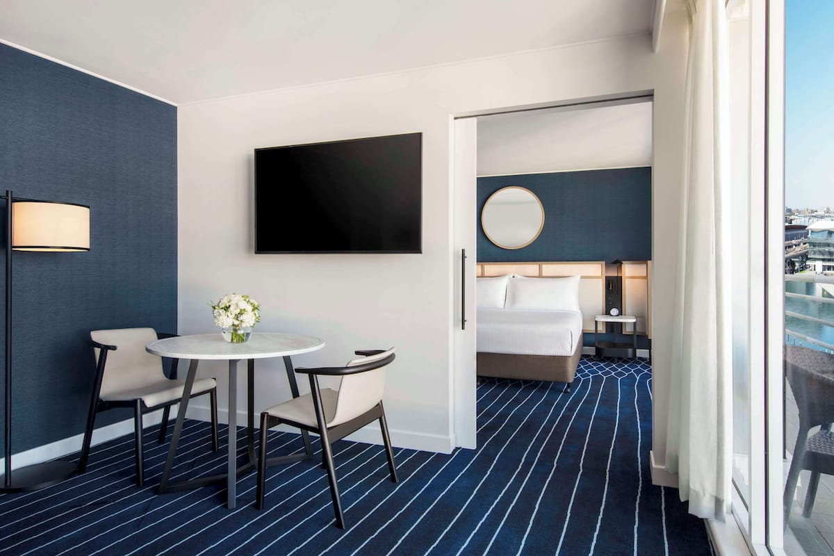 Una camera d'albergo con un letto, uno specchio rotondo, una TV, un tavolino con sedie e vista sulla città attraverso ampie finestre.