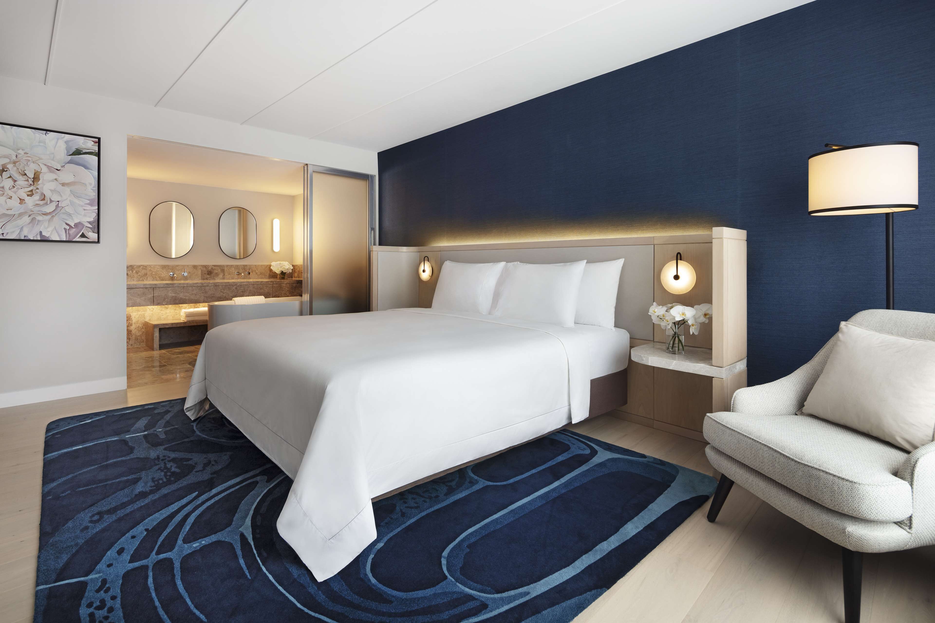 Una habitación de hotel moderna con una cama grande, una zona de estar con una silla, un tocador con espejo y una mesita de noche con lámpara.