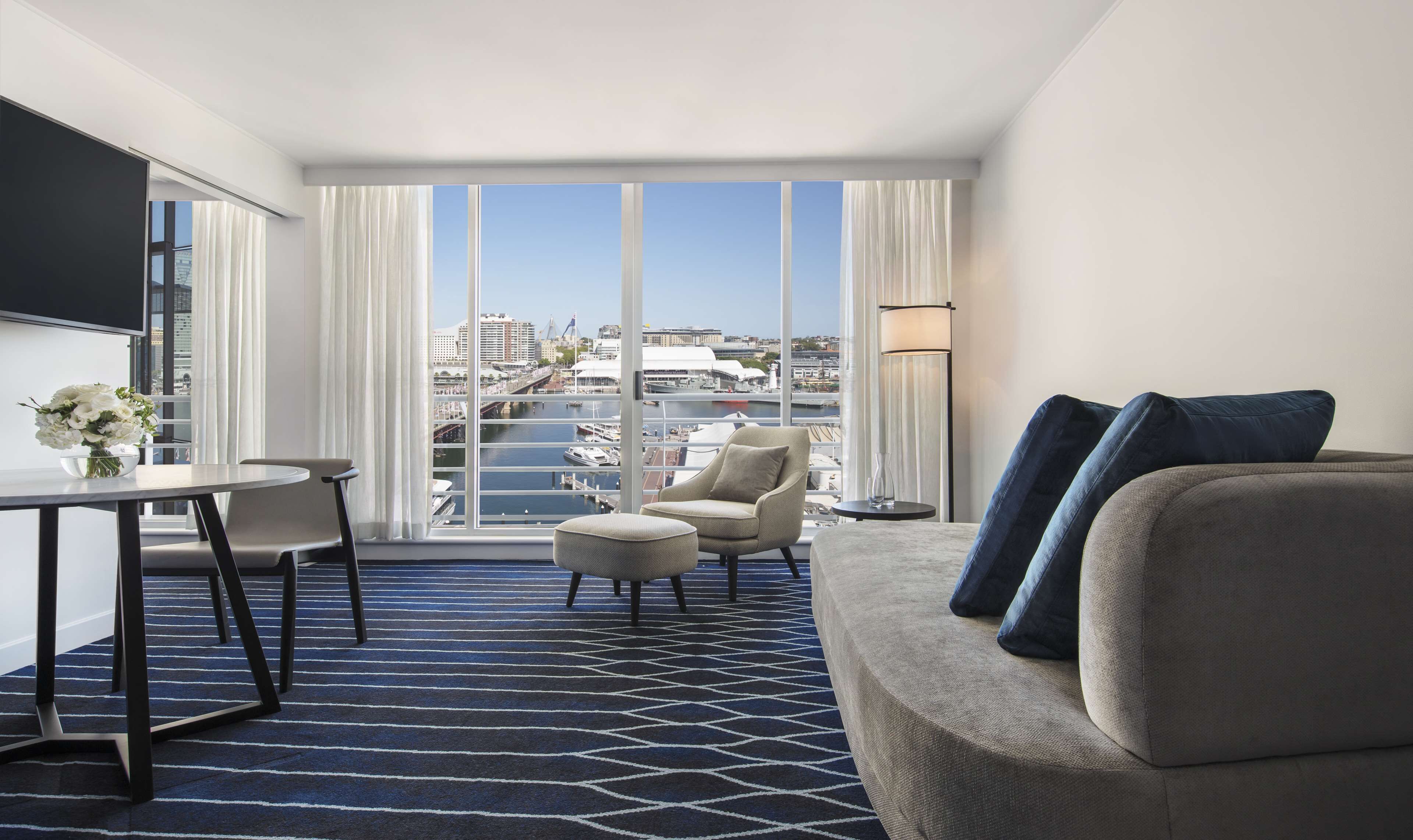 Habitación de hotel con un ventanal grande, un sofá, una silla, una mesa de comedor y un televisor.