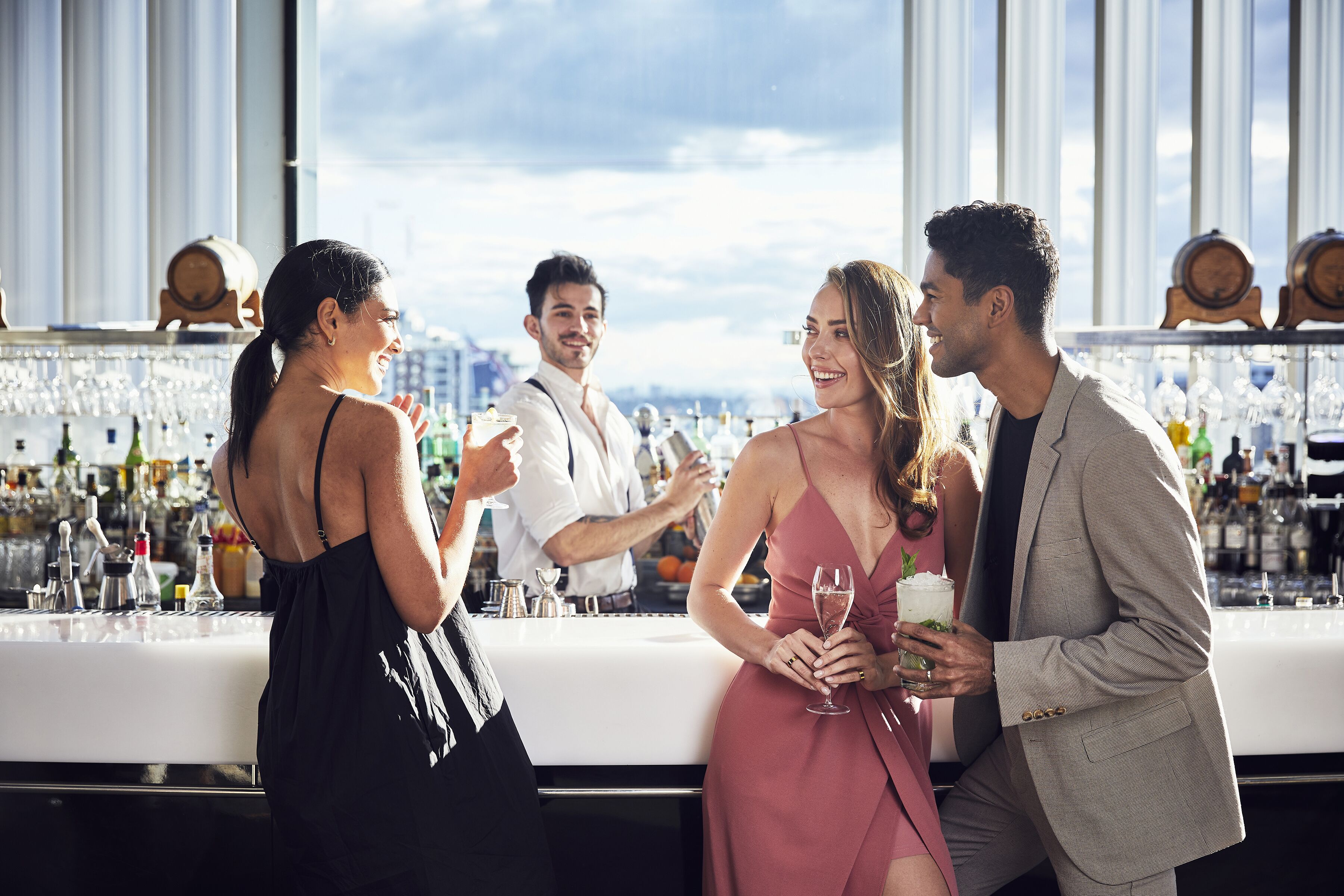 A bar area with a white countertop, bottles, glasses, and a city view through large windows.