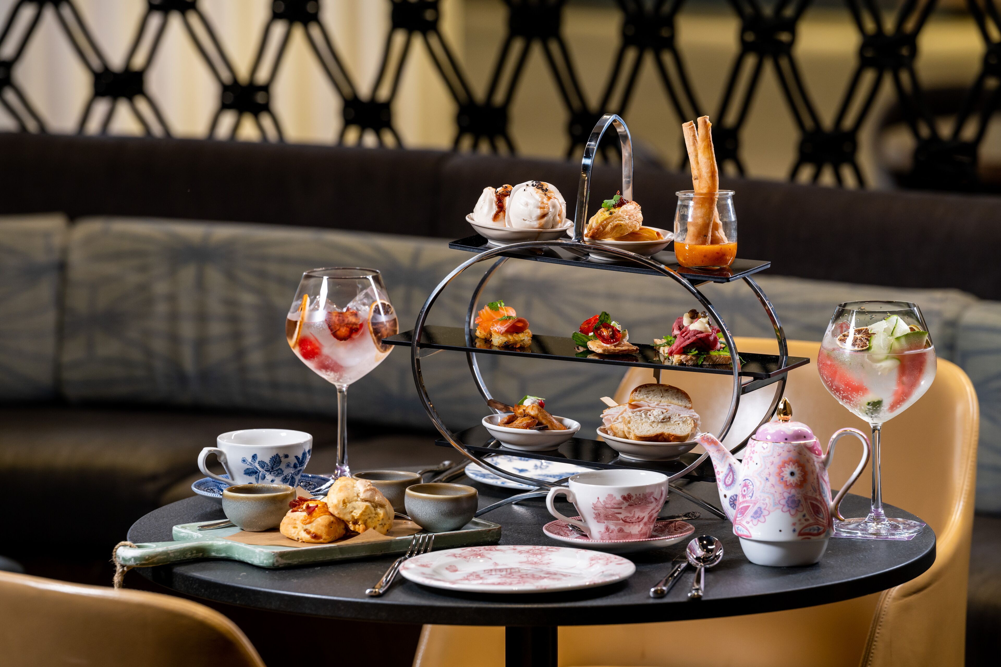 A three-tiered dessert stand with various pastries and desserts, a teapot, cups, and a glass of iced tea on a round table.