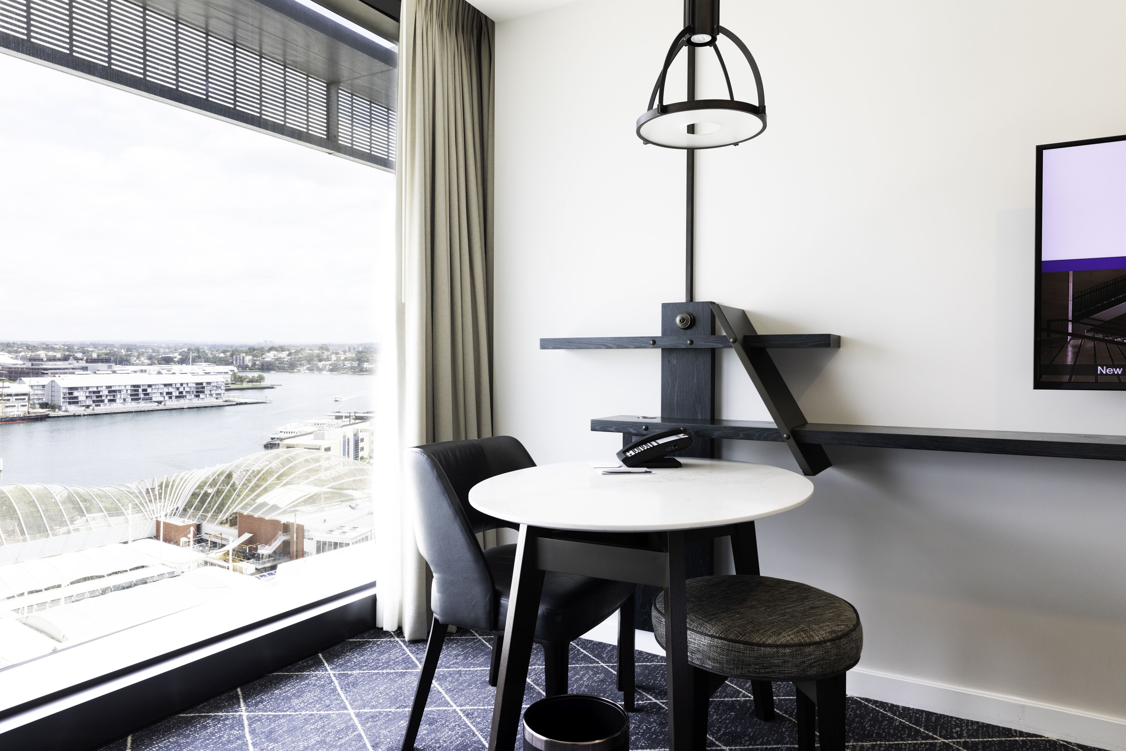 Una habitación de hotel moderna con una mesa redonda, dos sillas, un taburete pequeño, un televisor montado y vistas a la ciudad y al río.