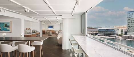 A modern bar area with a long counter and high stools, a kitchen in the background, and a view of a river and buildings.
