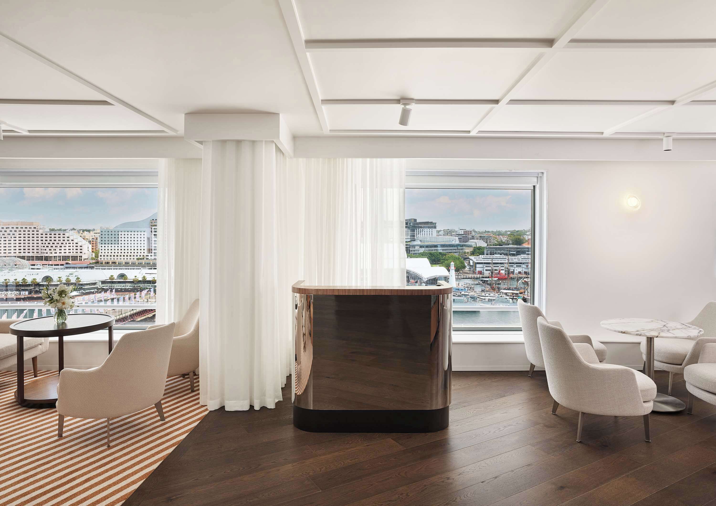 A modern hotel room with a large window offering a city view, a round wooden table, and two upholstered chairs.
