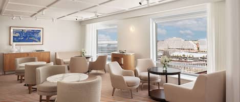 A modern hotel room with a large window offering a city view, a striped carpet, and several seating areas with a wooden cabinet and a painting on the wall.