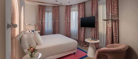 A hotel room with a bed, a TV, a small table, a chair, and curtains with a geometric pattern.