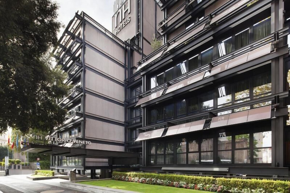 Um edifício moderno de hotel com um destaque para o letreiro 'NH Hotels', vários andares e um jardim frontal bem cuidado.