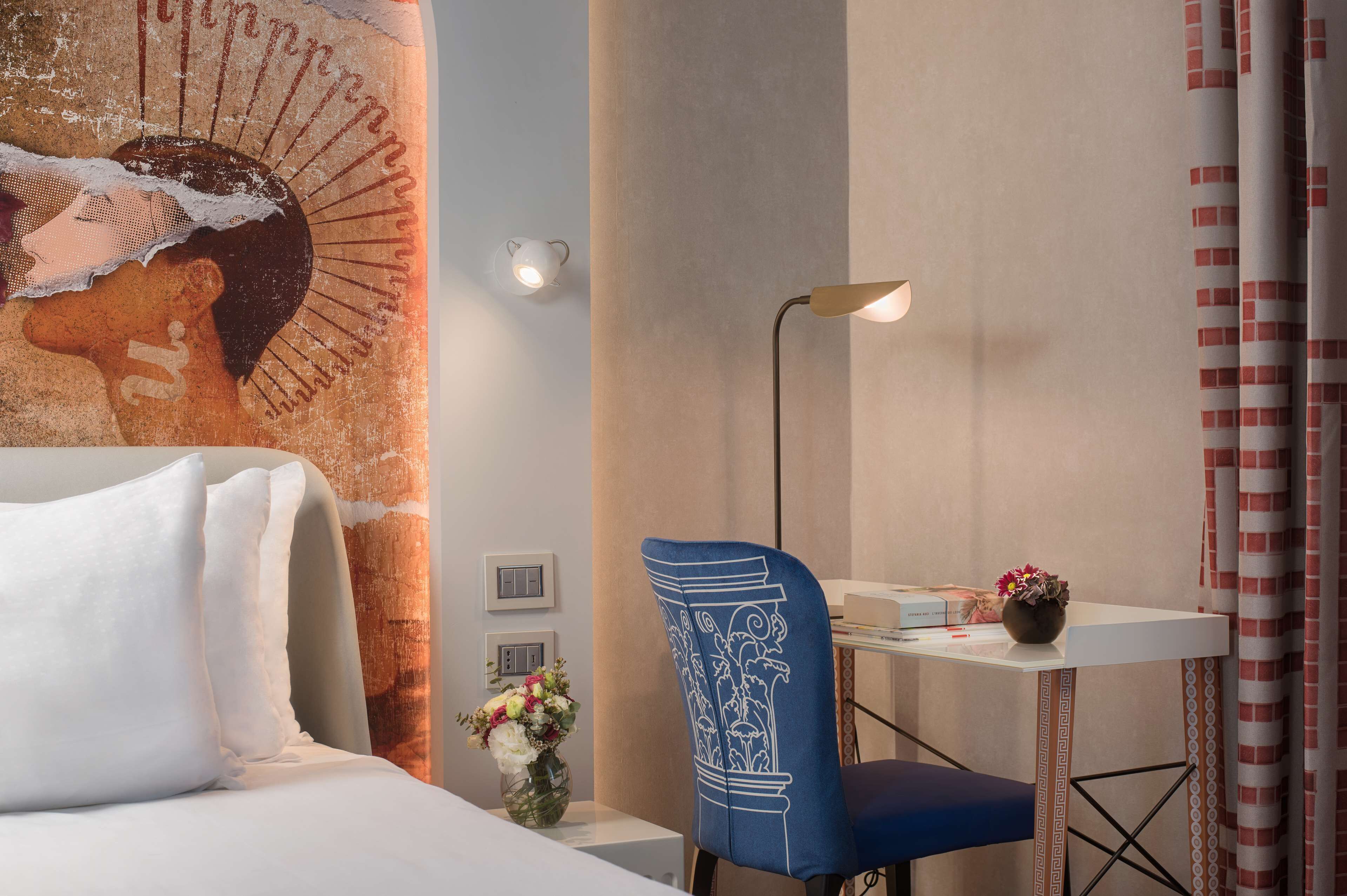 A hotel room with a bed, a desk with a chair, a lamp, a vase with flowers, and a wall mural.