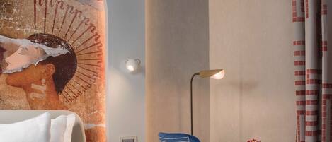 A hotel room with a bed, a desk with a chair, a lamp, a vase with flowers, and a wall mural.