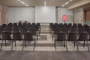 Egy konferenciaterem, melyben sorokban állnak a székek, szemben egy nagy képernyővel, egy emelvény és egy tábla található.
