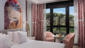 Une chambre d’hôtel avec un grand lit, deux fauteuils roses, une petite table avec un livre et un vase, et une vue sur les arbres à travers de grandes fenêtres.