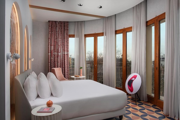 Quarto de hotel moderno com uma cama grande, uma mesa redonda pequena, uma cadeira e vista para o exterior por grandes janelas.