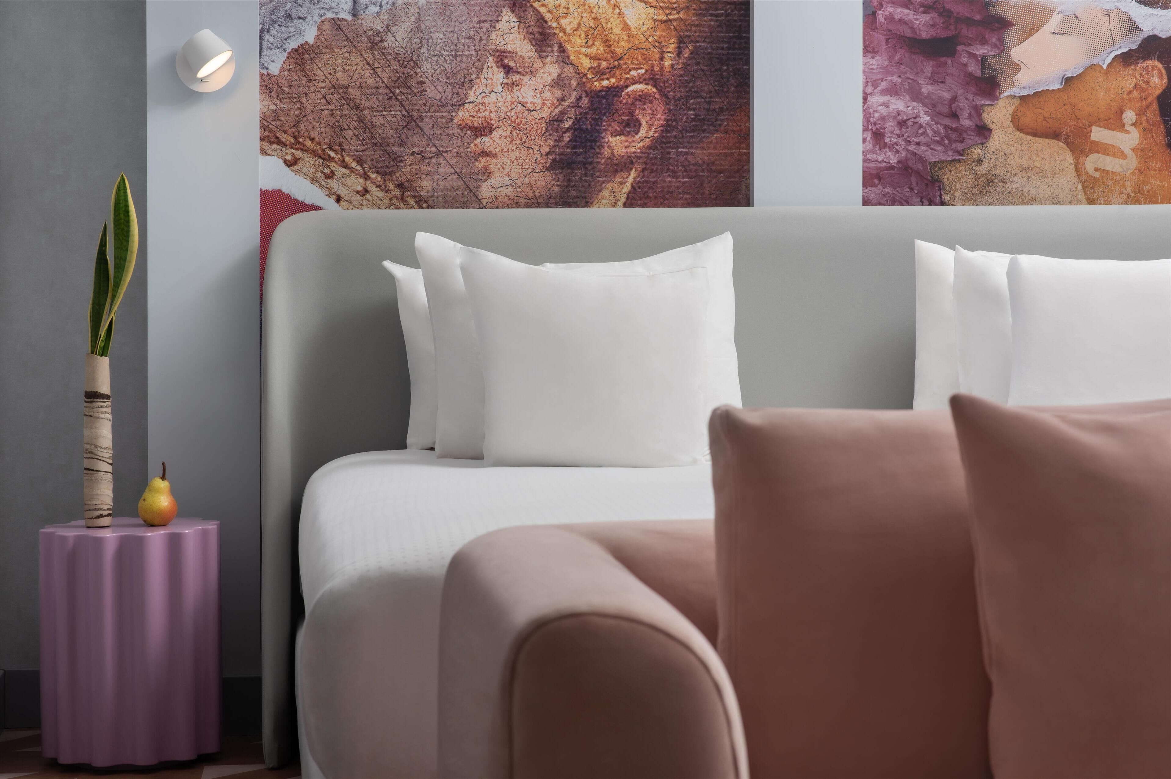 A modern bedroom with a grey headboard, white pillows, and a small purple side table with a plant and a fruit.
