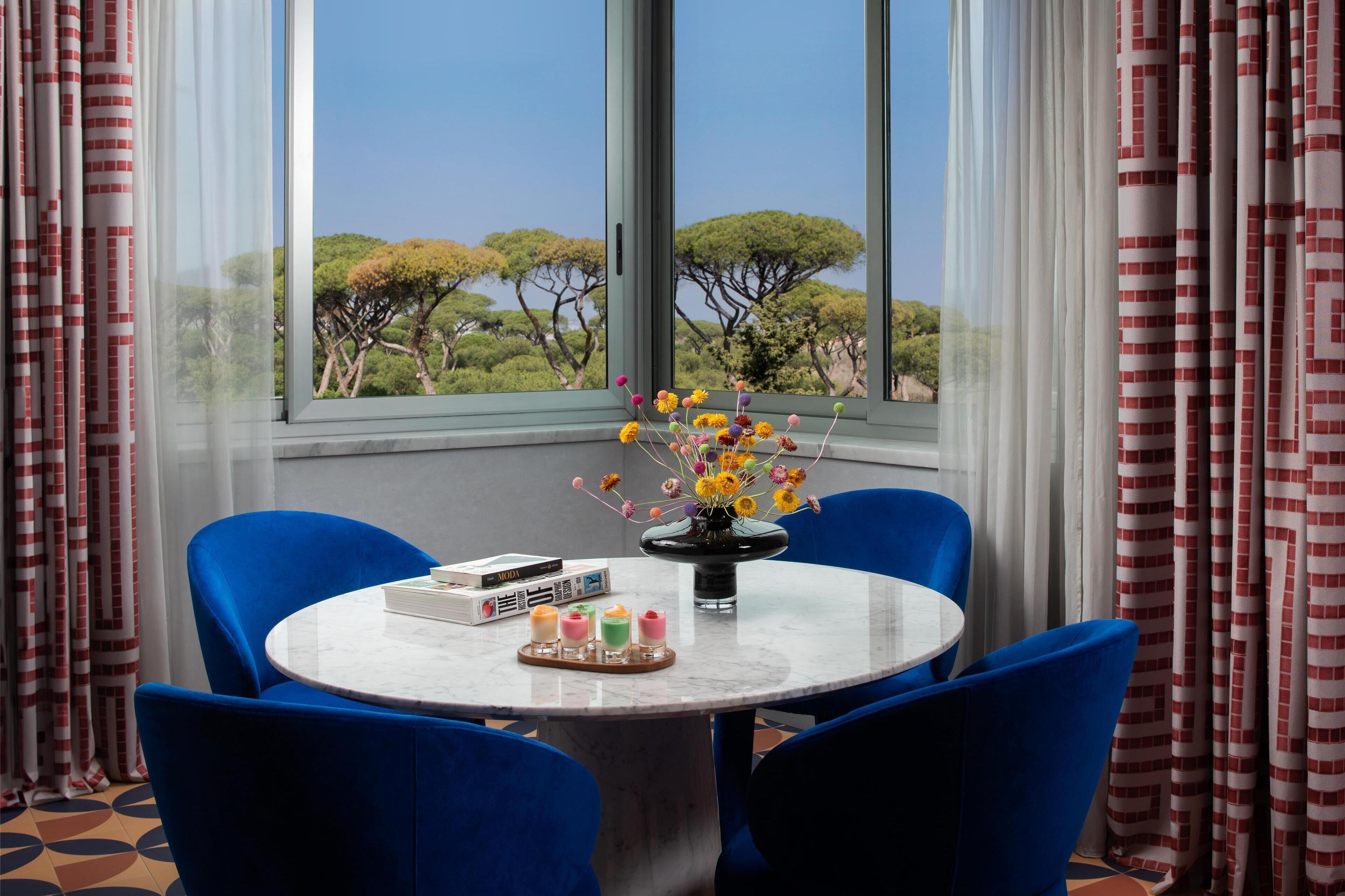 Una habitación con una mesa redonda, sillas azules, un jarrón con flores y vistas a árboles a través de amplias ventanas.