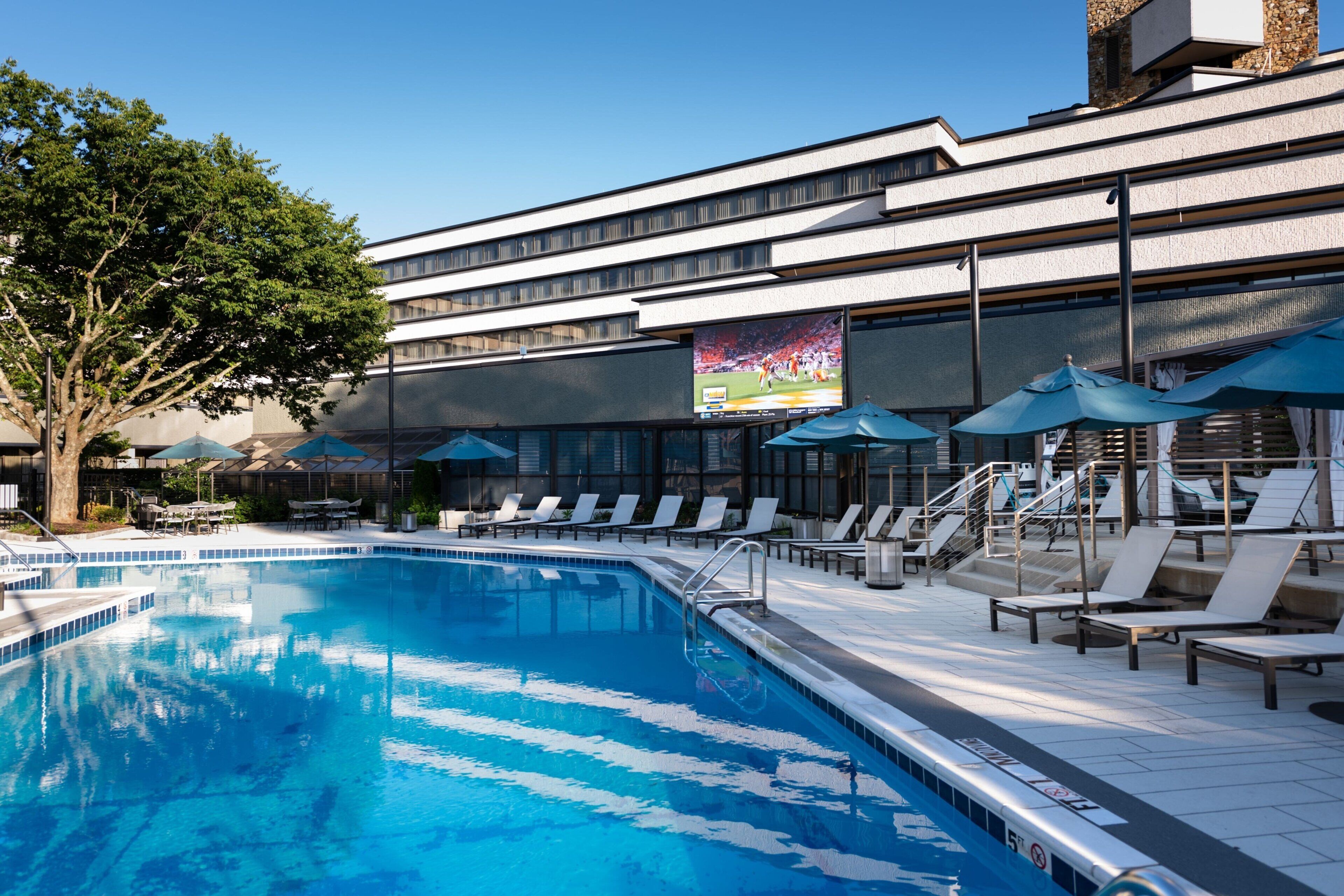 Ein Schwimmbecken mit Liegestühlen und Sonnenschirmen, einem großen Bildschirm und einem mehrstöckigen Gebäude.