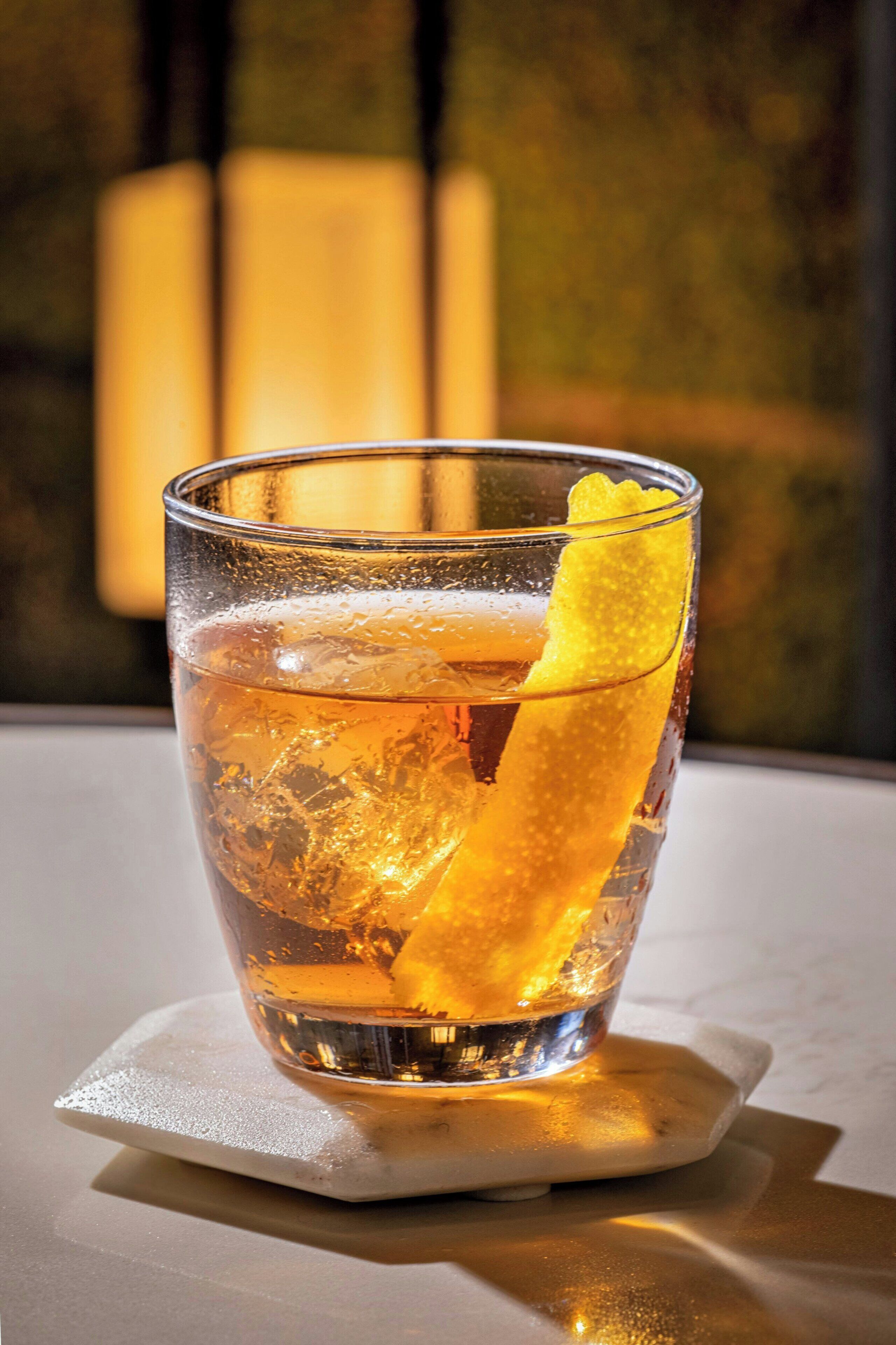 A glass of amber-colored beverage with a lemon slice garnish on a coaster.