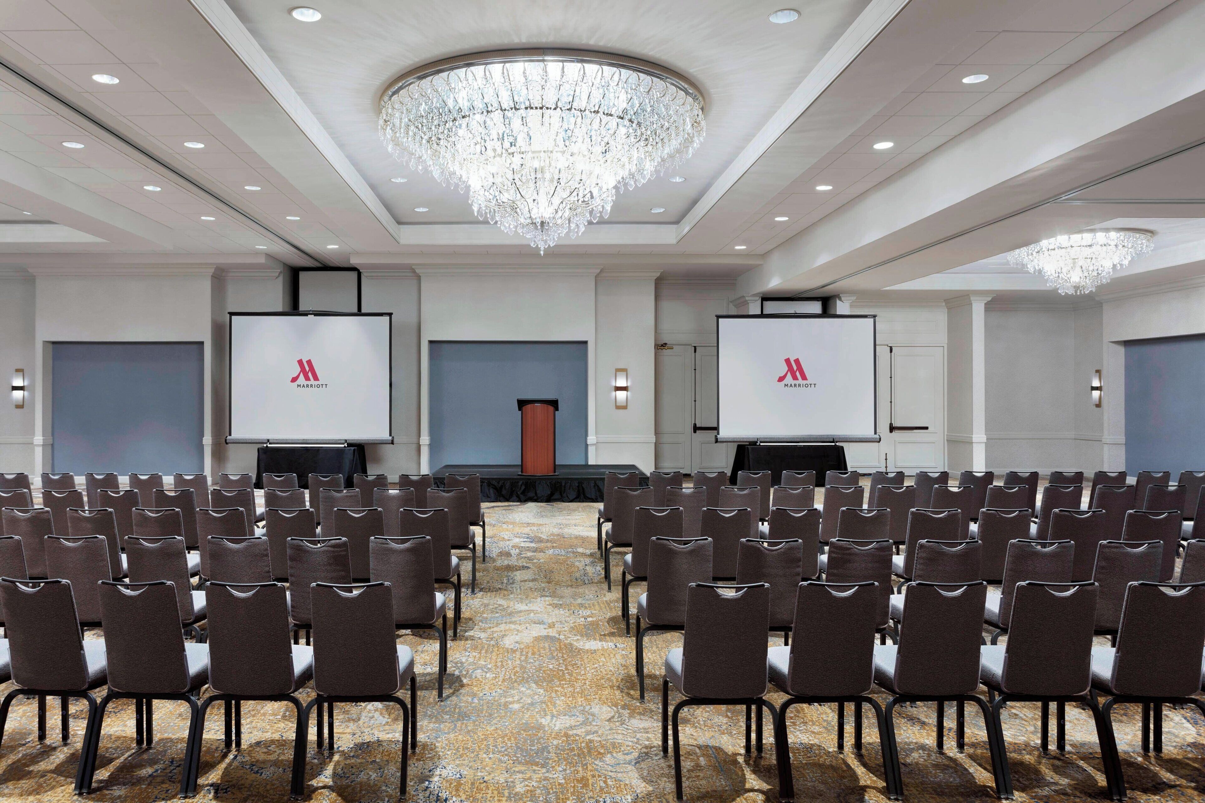 Ein Konferenzraum mit Reihen von Stühlen, einer großen Kronleuchter und zwei Bildschirmen, die das Marriott-Logo zeigen.