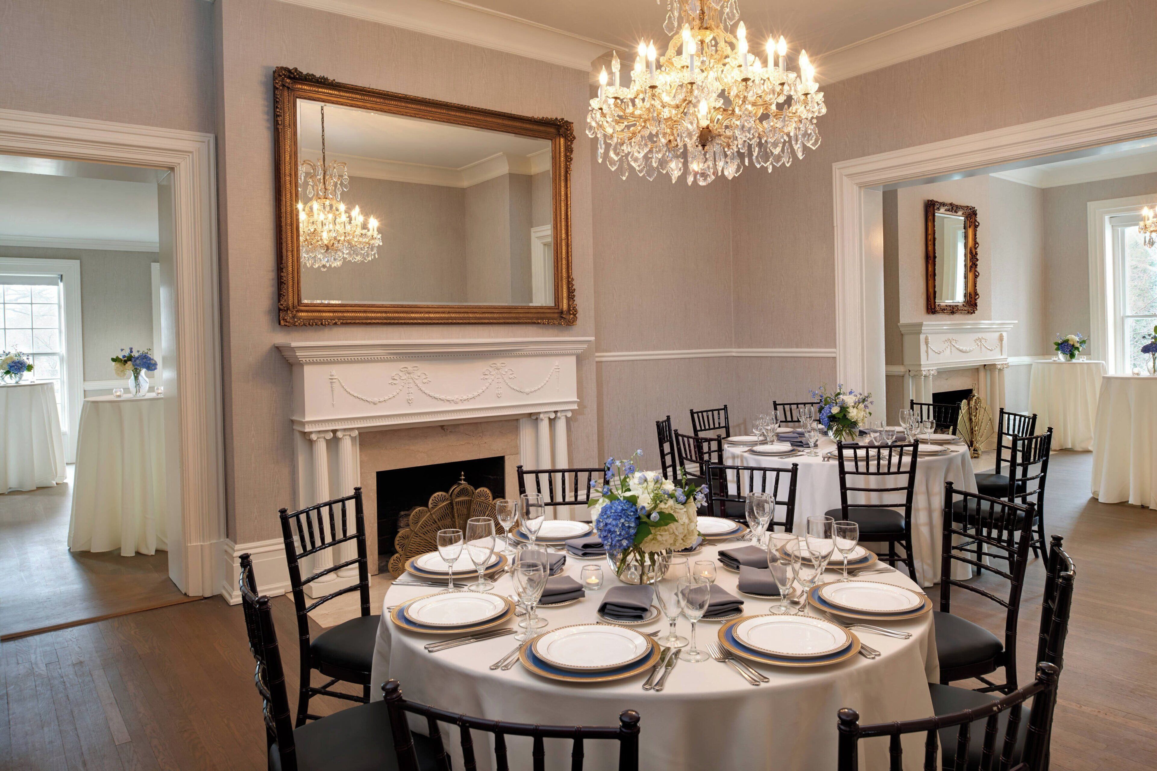 A formal dining room with round tables, black chairs, chandeliers, and a fireplace.